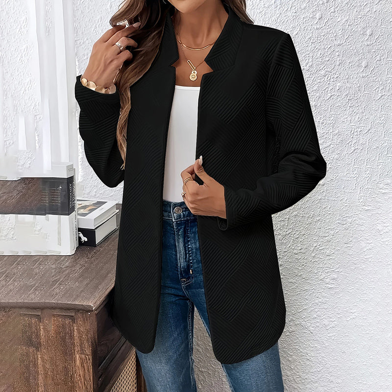 Person wearing a black blazer over a white top and blue jeans against a white wall.