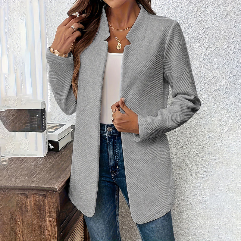 Woman wearing a gray cardigan over a white top and blue jeans against a white wall.