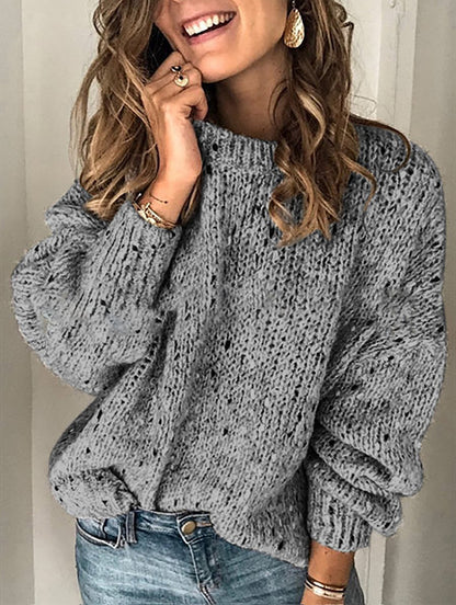 Woman wearing a gray knitted sweater and blue jeans, smiling.