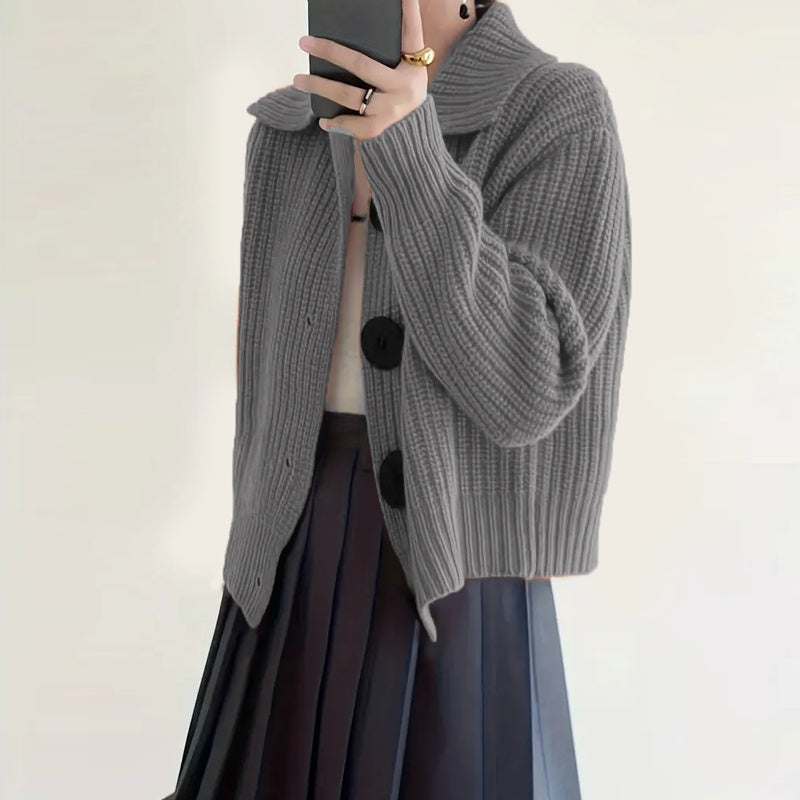 Person wearing a gray knitted cardigan with black pom-poms on a plain background