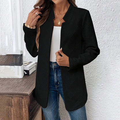 Person wearing a black blazer over a white top and blue jeans against a white wall.