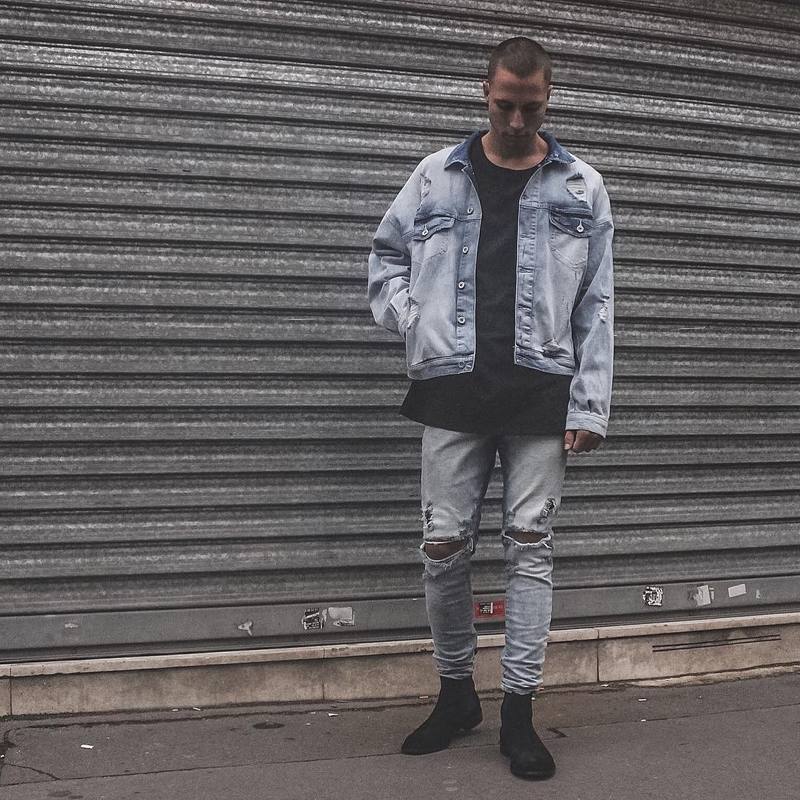 Person wearing a light blue denim jacket and jeans standing in front of a metal shutter.
