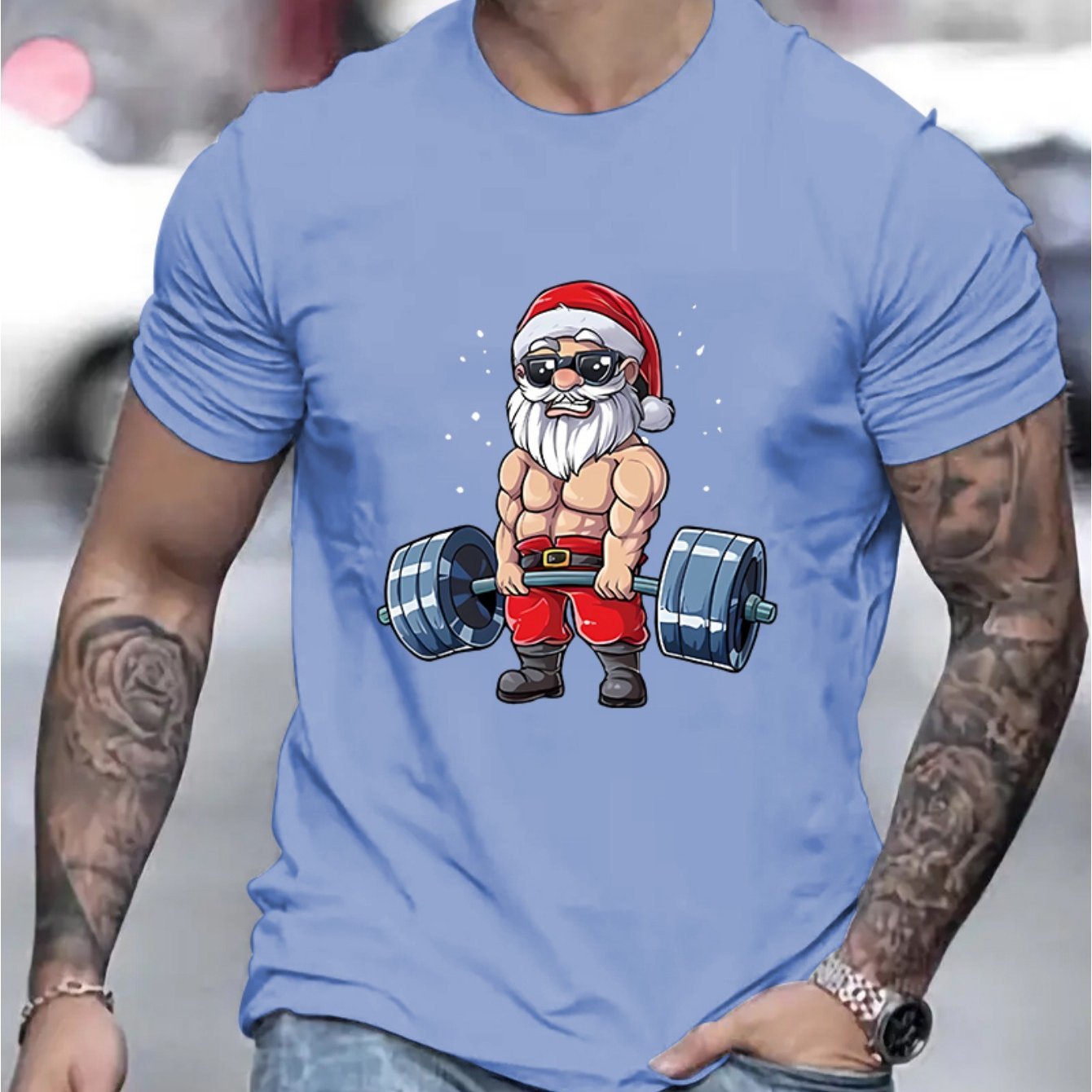 Light blue t-shirt with a graphic of a muscular Santa Claus lifting weights, worn by a person with tattoos.