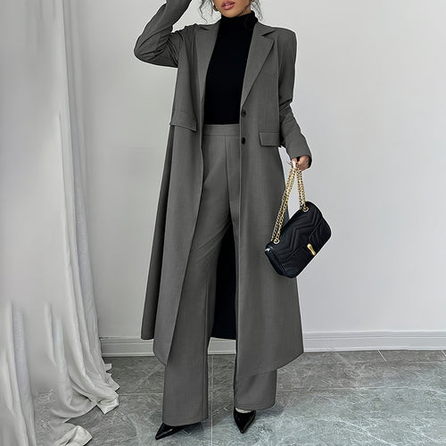 Person wearing a long gray coat and black pants, holding a black handbag against a plain background.