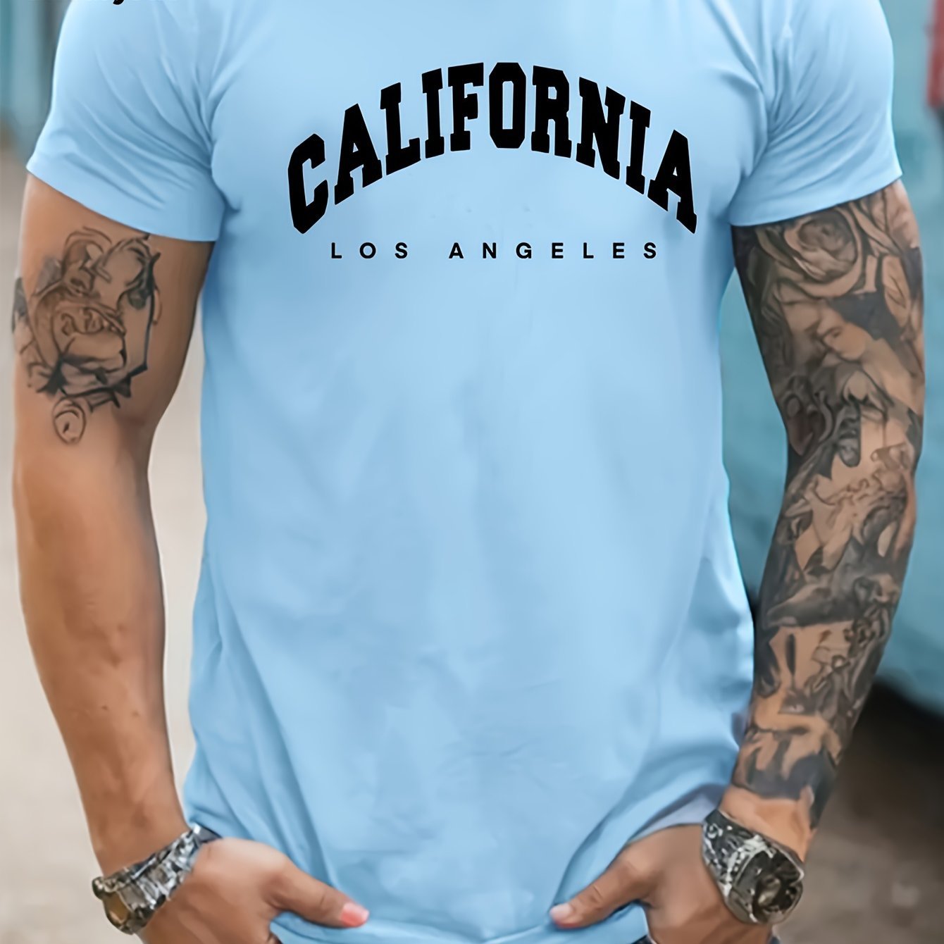 Person wearing a light blue t-shirt with 'California Los Angeles' text, tattoos on arms, and jewelry.